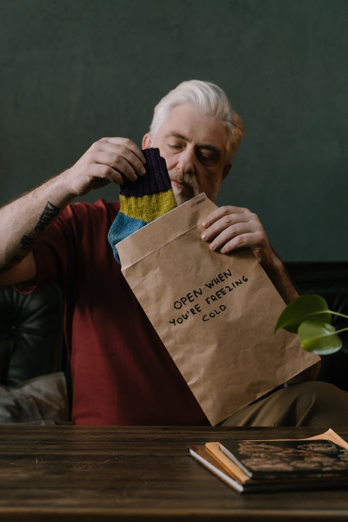 Senior man with beard opens a gift package labeled for cold weather, indoors.